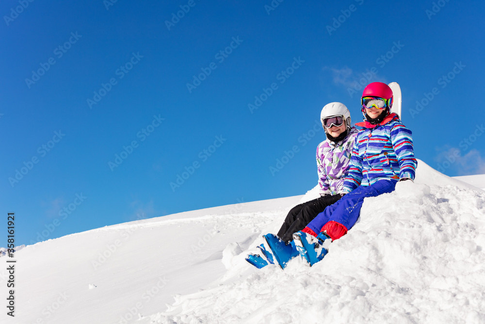 Two cute girls sit on pile of snow over blue background in sky outfit color mask and glasses on look at camera smiling
