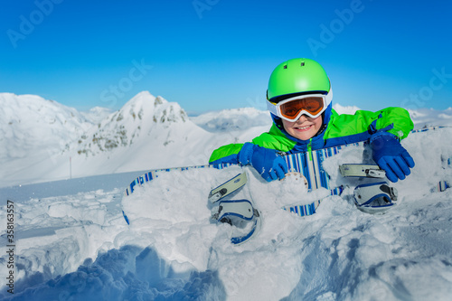 Little boy lay with snowboard in snow over mountains range wearing helmet and ski googles
