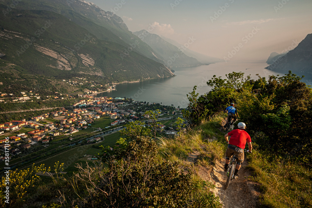 LAKE GARDA, ITALY. Two mountain bikers riding along a narrow single-track leading down towards the town of Riva del Garda. Evening light and mist over the lake.