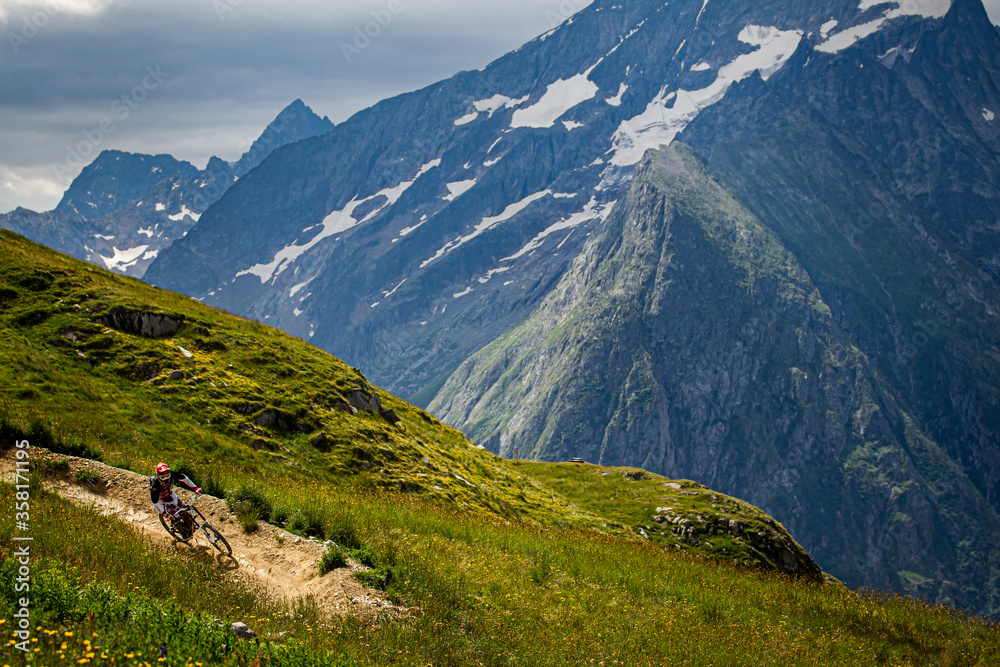 LES DEUX ALPES, FRANCE. A mountain biker riding a 'bermed' corner on a bike-park trail through grassy pasture, with patches of snow and dramatic alpine peaks behind.
