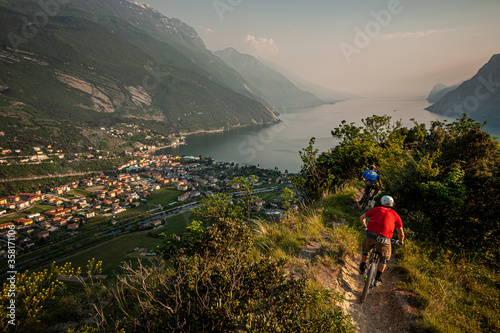 LAKE GARDA, ITALY. Two mountain bikers riding along a narrow single-track leading down towards the town of Riva del Garda. Evening light and mist over the lake. © Victor Lucas