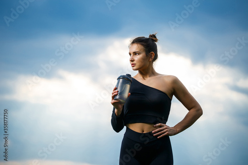 A girl of European appearance with a sports build in a black top and black leggings against a blue sky. She drinks water from a bottle. Healthy lifestyle, athletic brunette girl. Motivation for sports