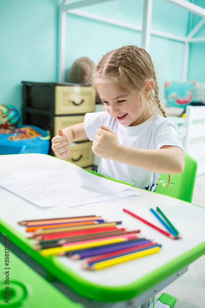 Little girl draws while sitting at a table in a room against the background of the wall Little girl draws while sitting at a table in a room against the background of the wall