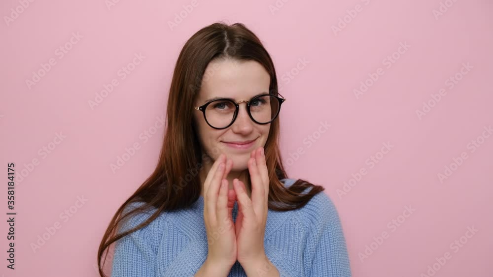 Portrait of young woman in eyeglasses schemes something, steepled fingers and looks with cunning expression aside, brunette girl smiles sly, wears blue sweater, poses over pink studio background Portrait of young woman in eyeglasses schemes something, steepled fingers and looks with cunning expression aside, brunette girl smiles sly, wears blue sweater, poses over pink studio background