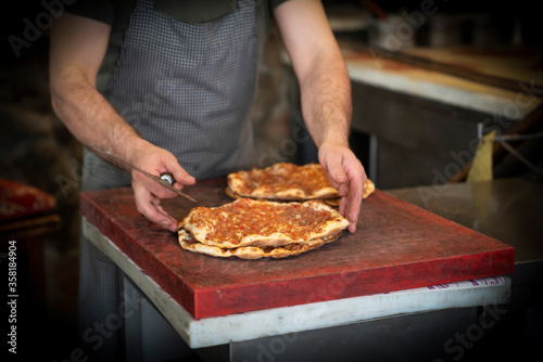 Baked lahmacun is a round, thin piece of dough topped with minced meat (most commonly beef or lamb), minced vegetables and herbs. Man cuts lahmacun.
