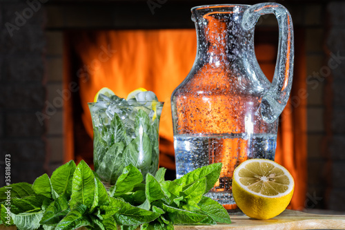 Lemonade with mineral water, lemon ,mint and ice in a jar and a glass before cozy fireplace.