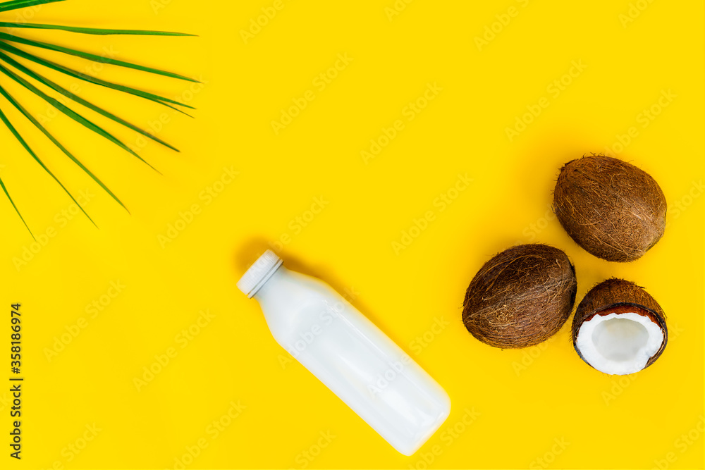 Top view bottle of fresh raw vegan coconut milk and fresh coconuts with a green palm leaf on the yellow background. Summer exotic refreshment. Natural plant based food. Copy space.