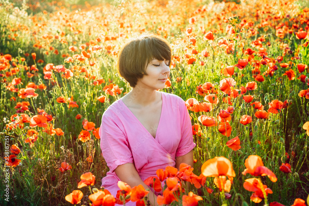 A happy young woman in a pink dress with closed eyes, relaxing in red poppies flowers meadow in sunset light. A simple pleasure for mental health. Nature relaxation. Selective focus. Copy space. A happy young woman in a pink dress with closed eyes, relaxing in red poppies flowers meadow in sunset light. A simple pleasure for mental health. Nature relaxation. Selective focus. Copy space.