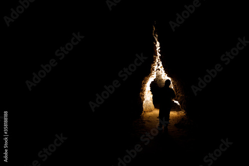 Man walking and exploring dark cave with light headlamp underground. Mysterious deep dark, explorer discovering mystery moody tunnel looking on rock wall inside.