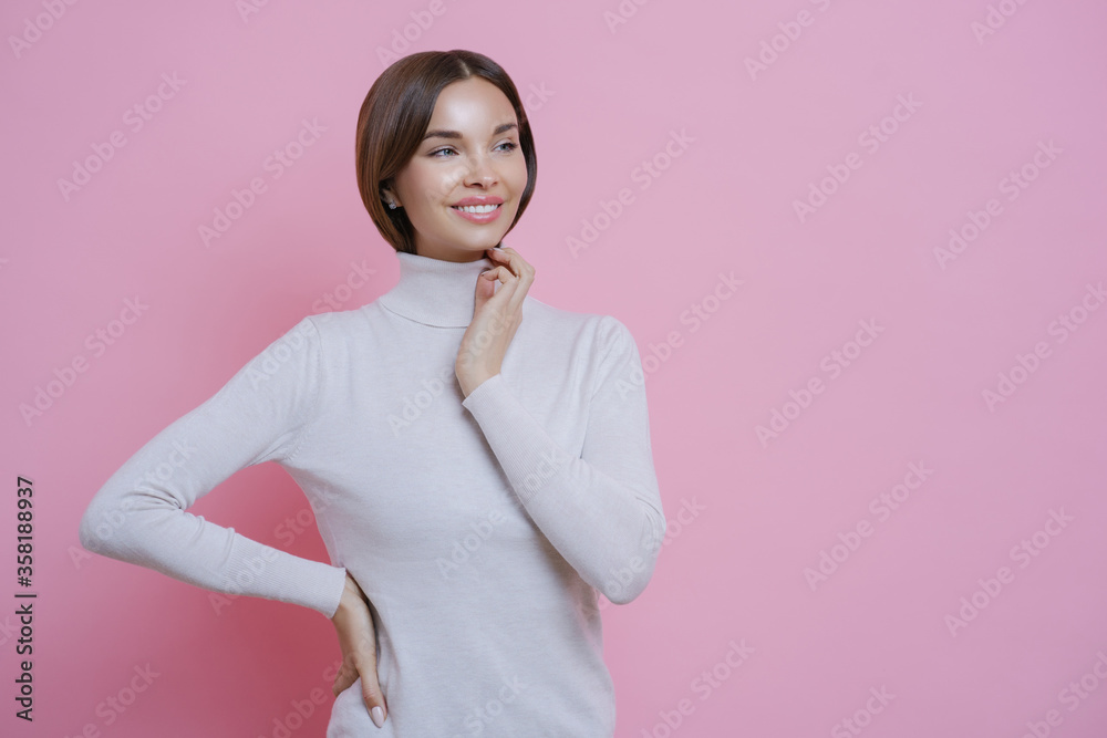 Beautiful tender young woman with toothy smile, looks aside pensively, dressed in casual white turtleneck, being in good mood, stands against pink background, blank free space for your promotion Beautiful tender young woman with toothy smile, looks aside pensively, dressed in casual white turtleneck, being in good mood, stands against pink background, blank free space for your promotion
