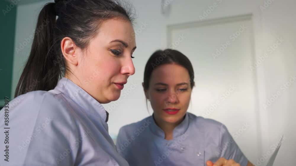 Two Young Beautiful Female Doctors Smiles and Discussing Treatment Plan Sitting Near a Computer in a Medical Office. Healthcare and Medicine Concept Two Young Beautiful Female Doctors Smiles and Discussing Treatment Plan Sitting Near a Computer in a Medical Office. Healthcare and Medicine Concept