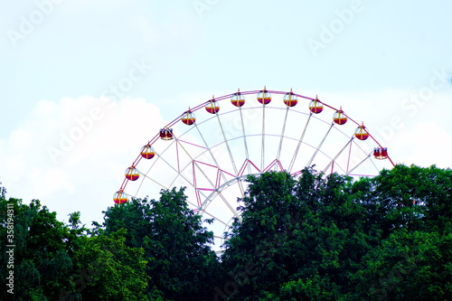Part of a large ferris wheel behind large trees against a blue sky