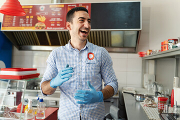 Young adult caucasian man entrepreneur standing at his fast food store in day smiling front view Chef in the kitchen having fun at work wearing protective medical gloves to prevent disease real people