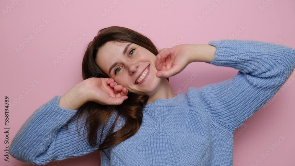 Lovely young woman with happy face expression, has sensual look, keeps hands near cheek, romantic expression, remembers something touching or pleasant, wears blue sweater, isolated on pink background Lovely young woman with happy face expression, has sensual look, keeps hands near cheek, romantic expression, remembers something touching or pleasant, wears blue sweater, isolated on pink background