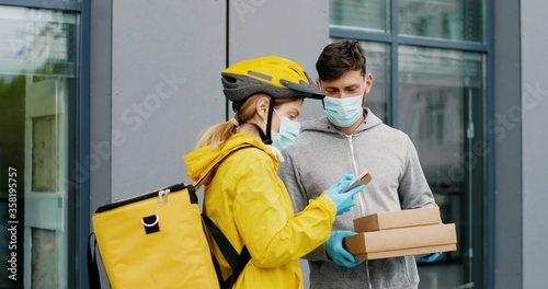 Caucasian young female courier in casque, gloves and medical mask giving carton parcels to man at street. Woman byciclist with bag bringing boxes to male customer outdoors. photo