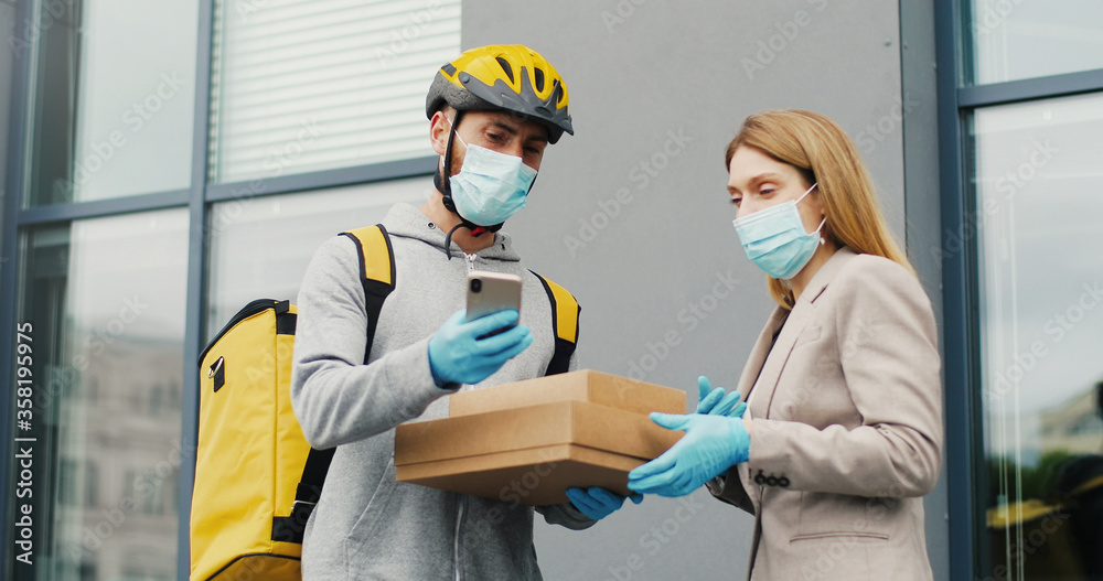 Young male courier in medical mask bringing carton boxes to woman on street and tapping on phone to check parcel. Food delivery service concept Young male courier in medical mask bringing carton boxes to woman on street and tapping on phone to check parcel. Food delivery service concept