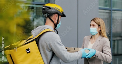 Portrait of delivery man with yellow thermal bag for food delivery in medical mask holding pizza boxes in front house and woman accepting a delivery of pizza boxes from courier.
