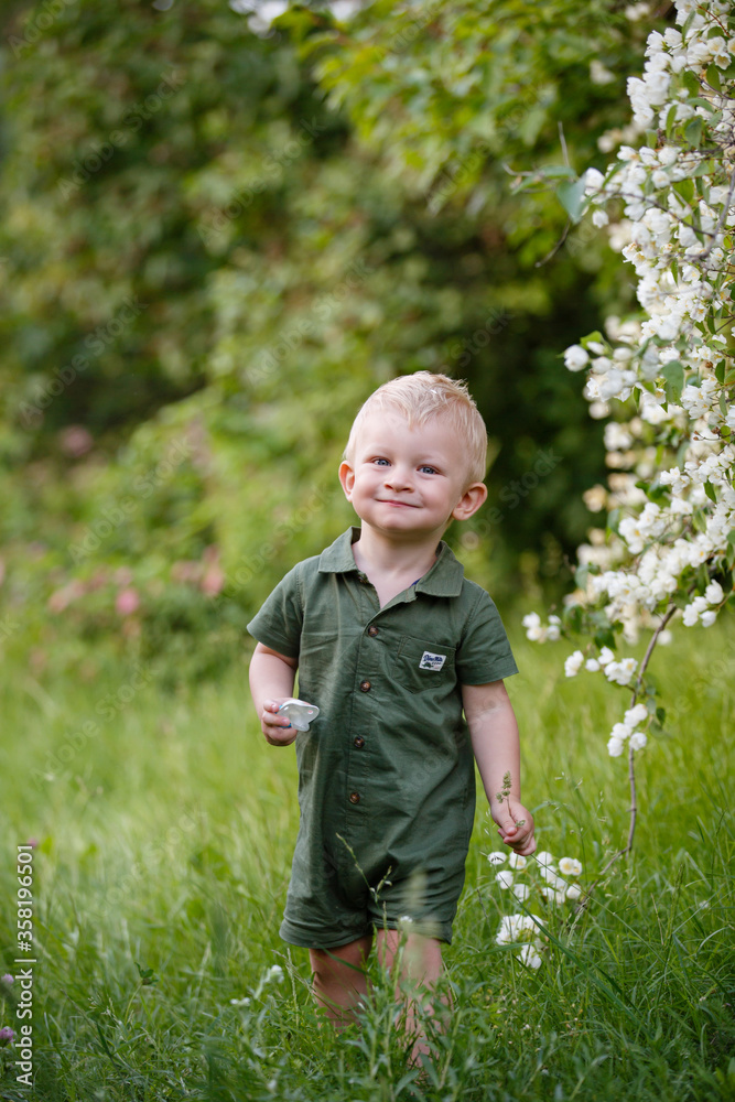 Little boy plays in blooming jasmine garden. Cute spring story. Toddler boy in green wreath in grass. Happy child in beautiful summer or spring sunny day Little boy plays in blooming jasmine garden. Cute spring story. Toddler boy in green wreath in grass. Happy child in beautiful summer or spring sunny day