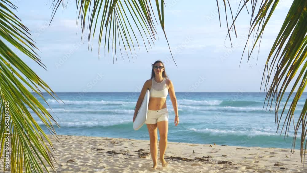 SLOW MOTION, PORTRAIT: Happy Caucasian woman walks away from the ocean with her surfboard. Fit female traveler on surfing trip in the Caribbean walks towards the camera after a day of awesome surfing. SLOW MOTION, PORTRAIT: Happy Caucasian woman walks away from the ocean with her surfboard. Fit female traveler on surfing trip in the Caribbean walks towards the camera after a day of awesome surfing.