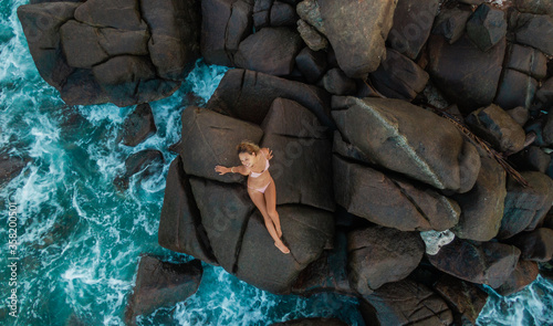 Beautiful slim blonde young woman in nude bikini sitting on rocks. Drone aerial shot. Picturesque ocean landscape with girl from above. Summer holiday in Sri Lanka photo