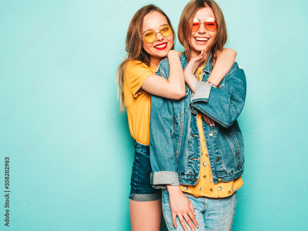 Two young beautiful smiling hipster girls in trendy summer yellow T-shirts and jeans jacket. Sexy carefree women posing near blue wall. Trendy and positive models having fun Two young beautiful smiling hipster girls in trendy summer yellow T-shirts and jeans jacket. Sexy carefree women posing near blue wall. Trendy and positive models having fun