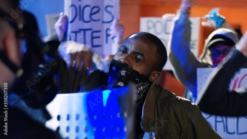 Street mess at night when male protesters fighting and quarreling with police in fully equipped costumes. African American man shouting at cop when standing at riot or demonstration. Aggressive guy. photo