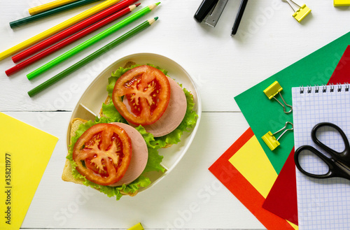 School supplies and lunch box with sausage sandwiches on a white wooden background