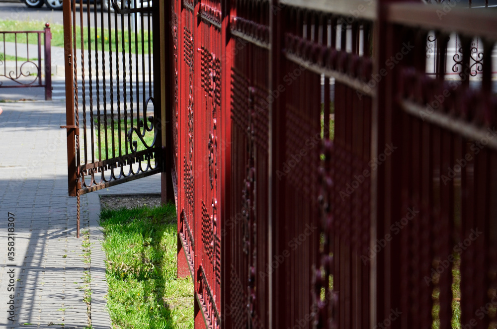 A metal fence and an open gate. Red fencing next to the pedestrian road. A metal fence and an open gate. Red fencing next to the pedestrian road.