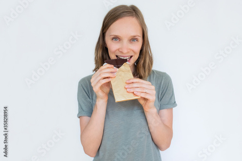 Beautiful woman biting chocolate bar in gold foil, smiling and looking at camera. Portrait of young lady isolated on white background. Front view. Tasty food and sweets concept