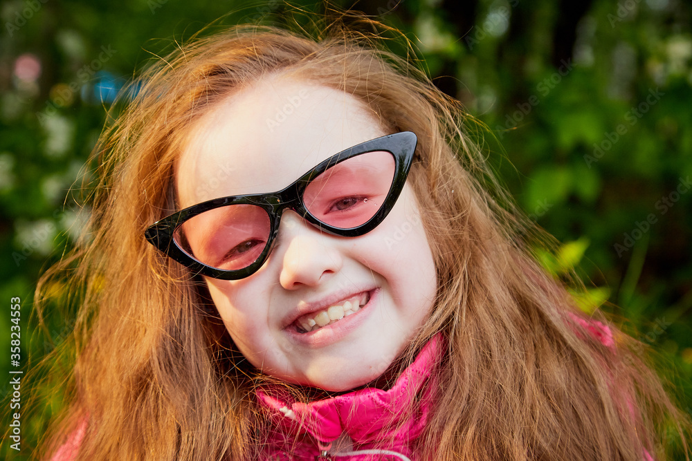 Pretty cute little girl with long hair and in pink glasses posing near a blooming apple tree with white flowers in a summer park Pretty cute little girl with long hair and in pink glasses posing near a blooming apple tree with white flowers in a summer park