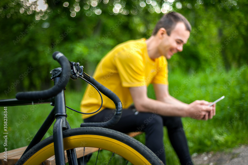 Obraz premium A young man sitting on a bench in a public Park communicates via mobile communication. In the background with his bike. Among trees and vegetation.