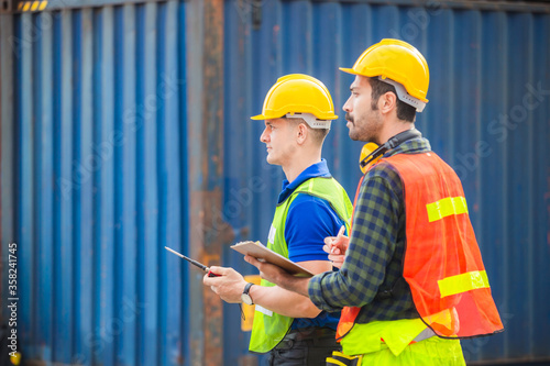 Engineer man in hardhat and safety vest talks on two-way radio, foreman worker holding clipboard checklist at containers cargo