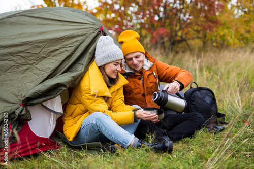 trekking and hiking concept - portrait of couple sitting near green tent and drinking tea in autumn forest