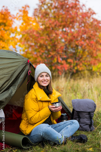 portrait of woman sitting near green tent and drinking tea in autumn forest