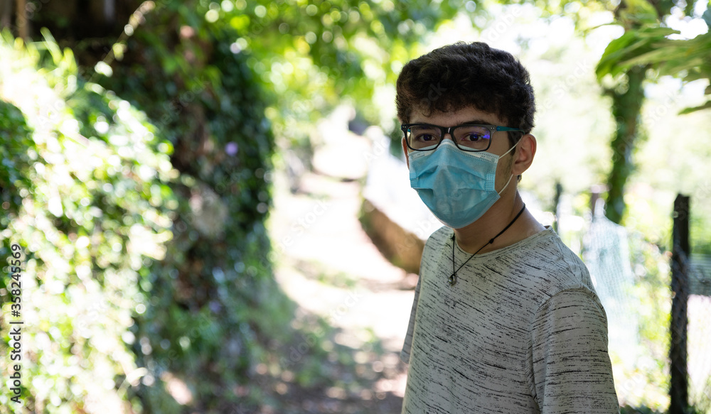 Portrait of a Caucasian boy wearing a mask to protect himself from the coronavirus during a field trip after the lockdown. In the background a path that creeps into the woods.