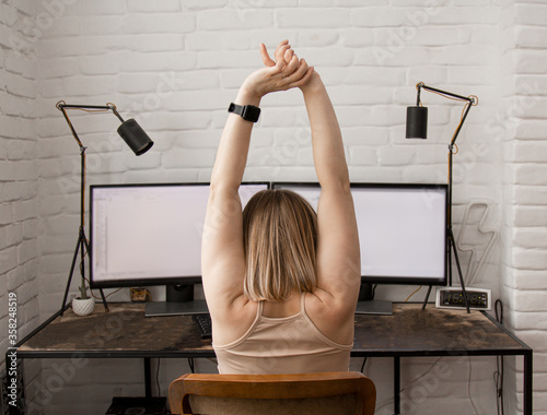a woman sits at a working table near a computer with two monitors takes a break flexing her neck suffers from pain caused by poor improper posture, sedentary work, sat for a long time at the compter