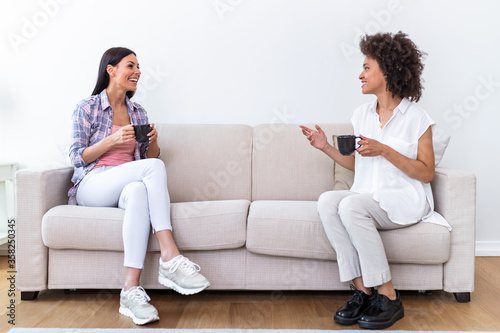 Two female friends in social distancing sitting on sofa. Best friends having coffee together while separated by social distancing on sofa at home