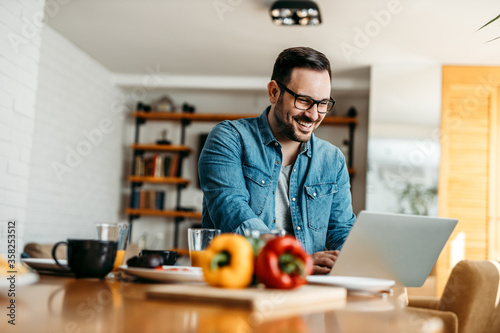 Portrait of a cheerful man using laptop at kitchen table in cozy apartment.