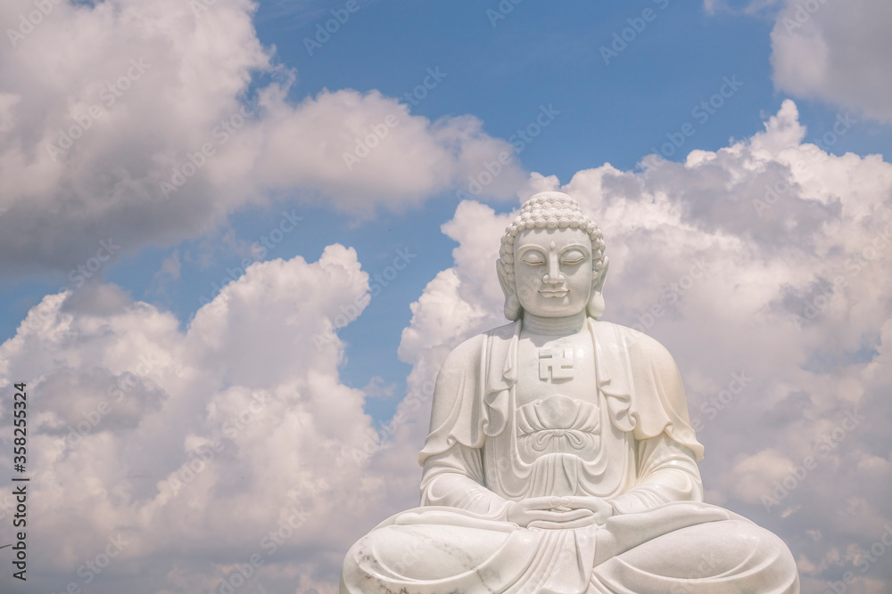 the solemn white statue of Buddha sitting on a high pedestal behind is a clear blue sky with white clouds