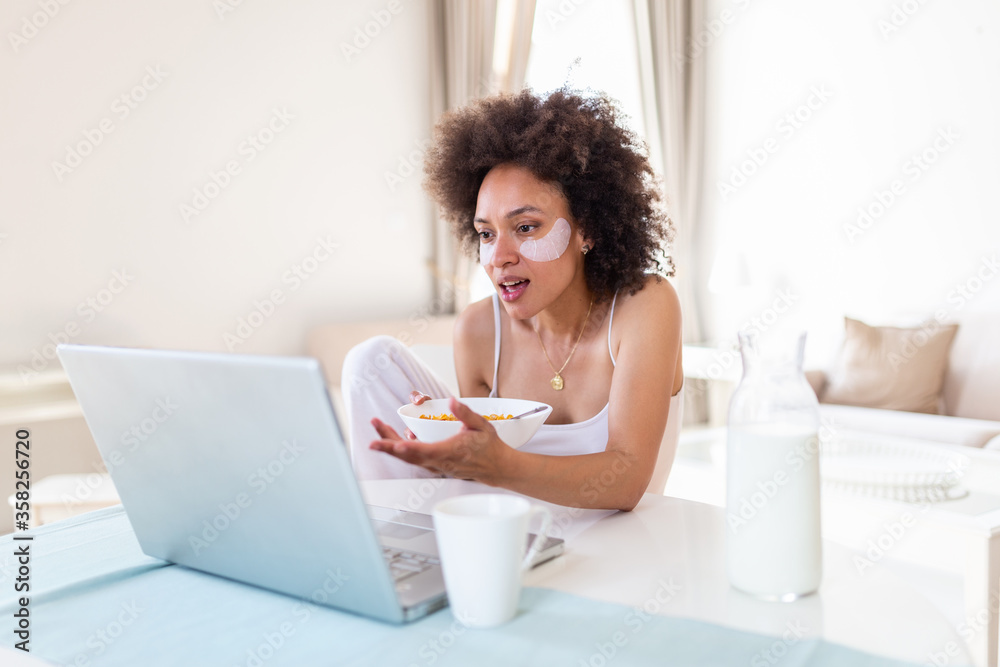 Image of happy young amazing woman sitting indoors at the table with laptop holding corn flakes. Looking at laptop computer and talking to her friends via video call. Image of happy young amazing woman sitting indoors at the table with laptop holding corn flakes. Looking at laptop computer and talking to her friends via video call.