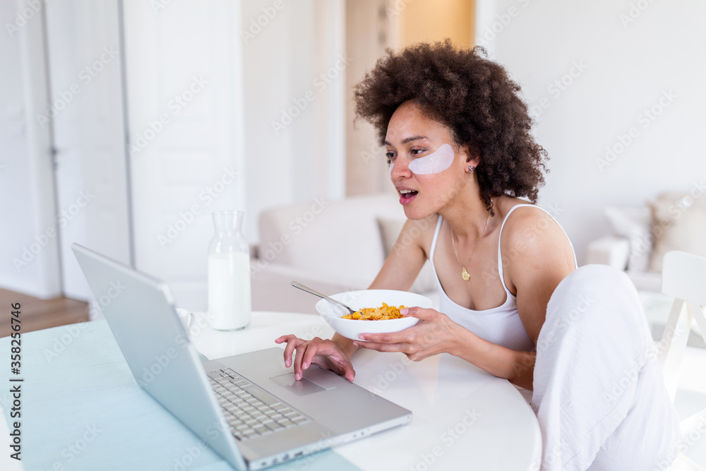 Attractive woman using her laptop computer while having breakfast and studying in home indoors. Freelance female model girl working on her laptop in the morning . Cup of milk with corn flakes