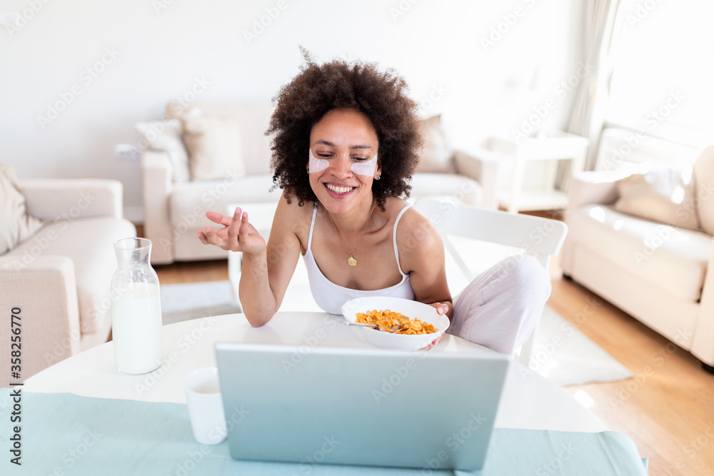 Image of happy young amazing woman sitting indoors at the table with laptop holding corn flakes. Looking at laptop computer and talking to her friends via video call. Image of happy young amazing woman sitting indoors at the table with laptop holding corn flakes. Looking at laptop computer and talking to her friends via video call.