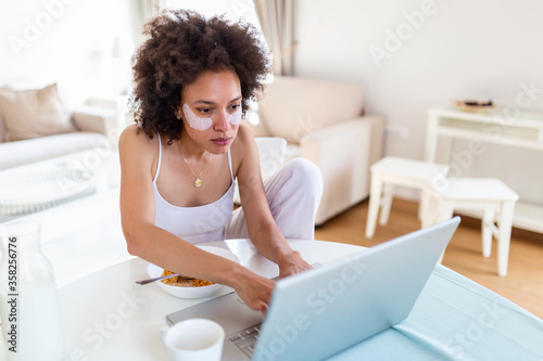 Beautiful young arfican american woman typing on laptop computer at home. Cheerful lady eating corn flakes and working on laptop at home