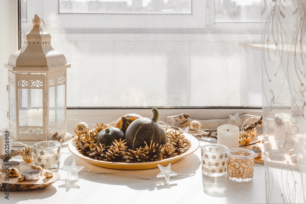 Pumpkins and cones on a golden tray and candles in glass glasses. Autumn window sill decoration. Autumn decor.