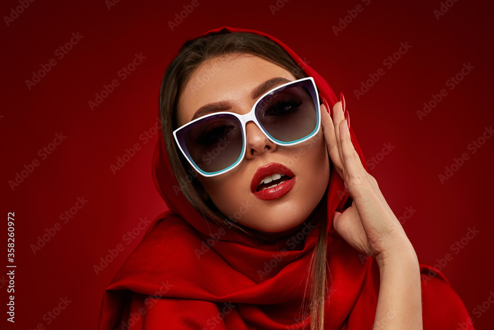 Portrait of chic brunette sexy girl with luxurious make-up in white sunglasses and red headscarf on red background