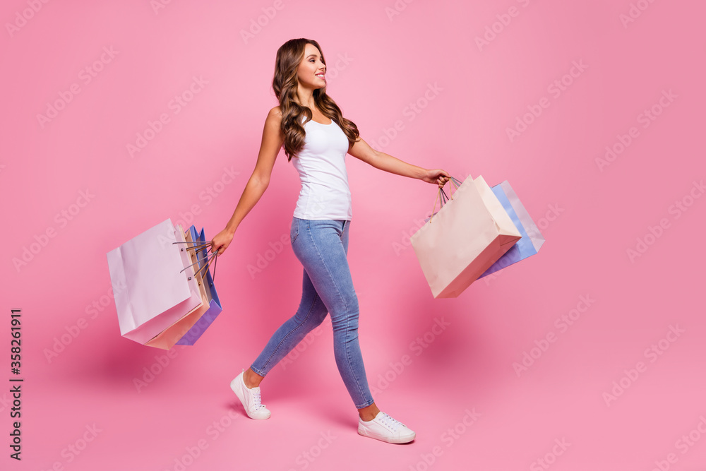 Full length profile photo of attractive lady carry many shopping packs addicted shopper walking store mall wear white casual tank-top jeans shoes isolated pink color background