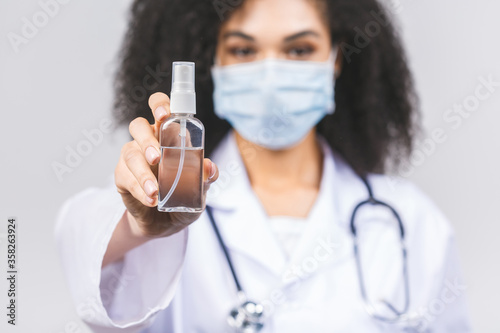 Young african american woman doctor in medical uniform and protective mask using hand sanitizer gel  isolated on grey background. Hand hygiene and antibacterial protection.