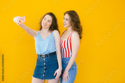 Two happy elegant women in dresses posing together and making selfie on smartphone over yellow background