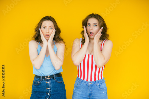Two shocked pretty women looking at the camera while holding their cheeks over yellow background