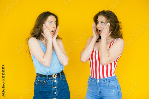 Two shocked pretty women looking at the camera while holding their cheeks over yellow background